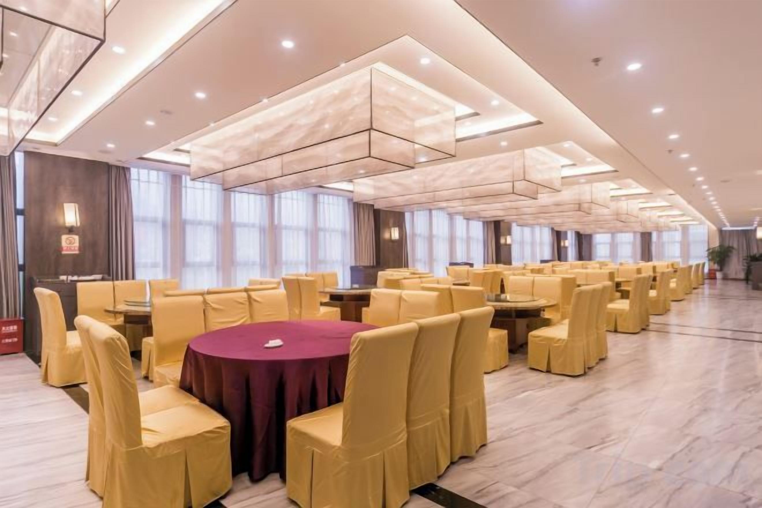 A spacious banquet hall with round tables and yellow chairs, large windows, and a modern ceiling design.