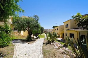 Un sendero adoquinado atraviesa un patio con edificios a ambos lados, rodeado de follaje y un cielo despejado.