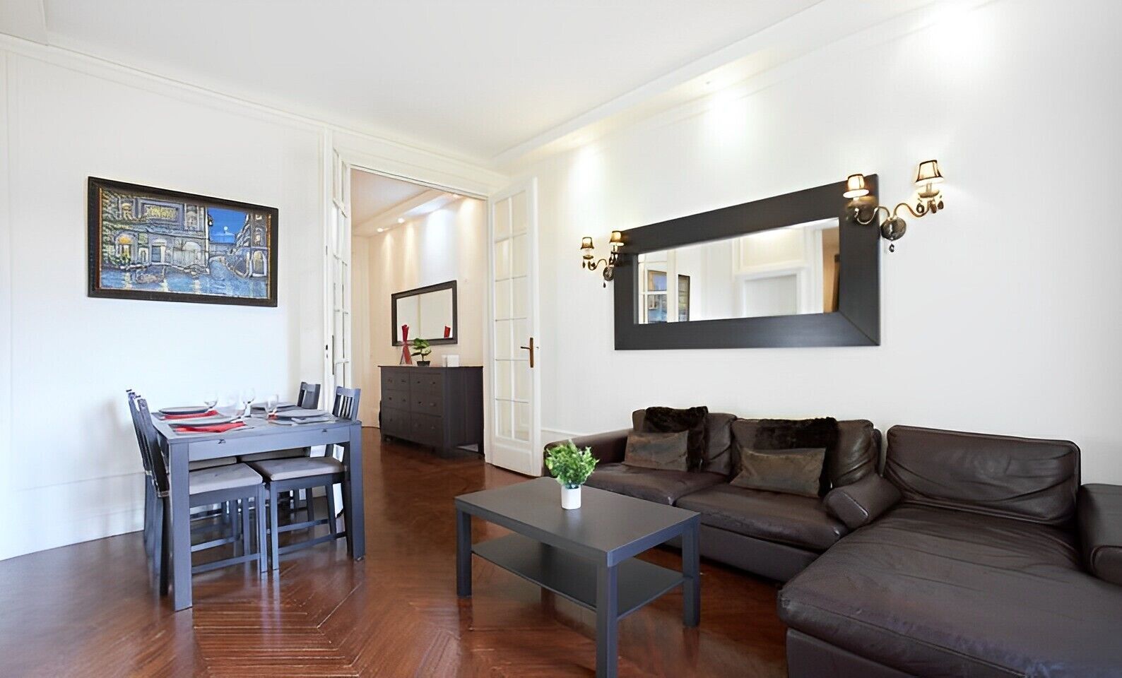 A modern living room with a dark brown leather sofa, a dining table with chairs, a coffee table with a plant, and a large mirror on the wall.