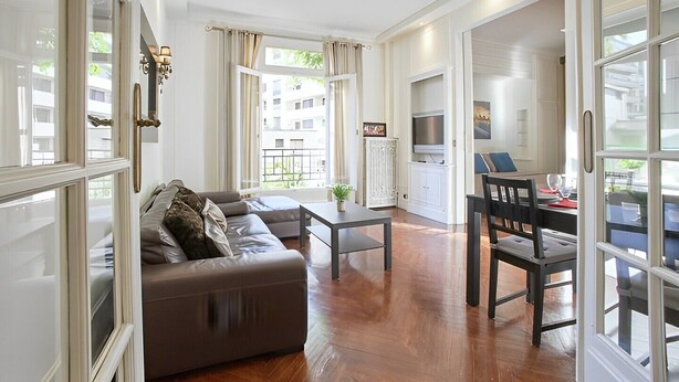 Un salon moderne comprenant un canapé, une table à manger et des chaises. Il y a également un coin cuisine avec un téléviseur et des placards. De grandes fenêtres laissent entrer la lumière naturelle et offrent une vue sur l’extérieur.