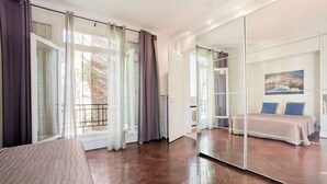 Une chambre dotée d’un grand miroir, d’un plancher en bois, d’un lit avec des oreillers bleus et offrant une vue sur le paysage urbain par la fenêtre.