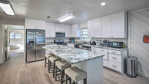 A modern kitchen with white cabinets, a stainless steel refrigerator, a microwave, a stove, and a sink. There are bar stools and a trash can. - Spacious Family Home w/ Backyard in Gainesville! (Gainesville)