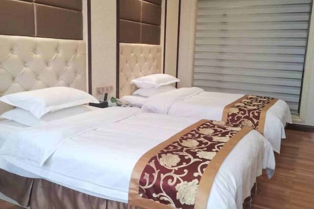 Une chambre d’hôtel avec deux lits, chacun recouvert d’une courtepointe blanche et agrémenté d’un chemin de table décoratif rouge et or. La chambre est dotée d’une tête de lit capitonnée et d’une fenêtre fermée avec des stores.