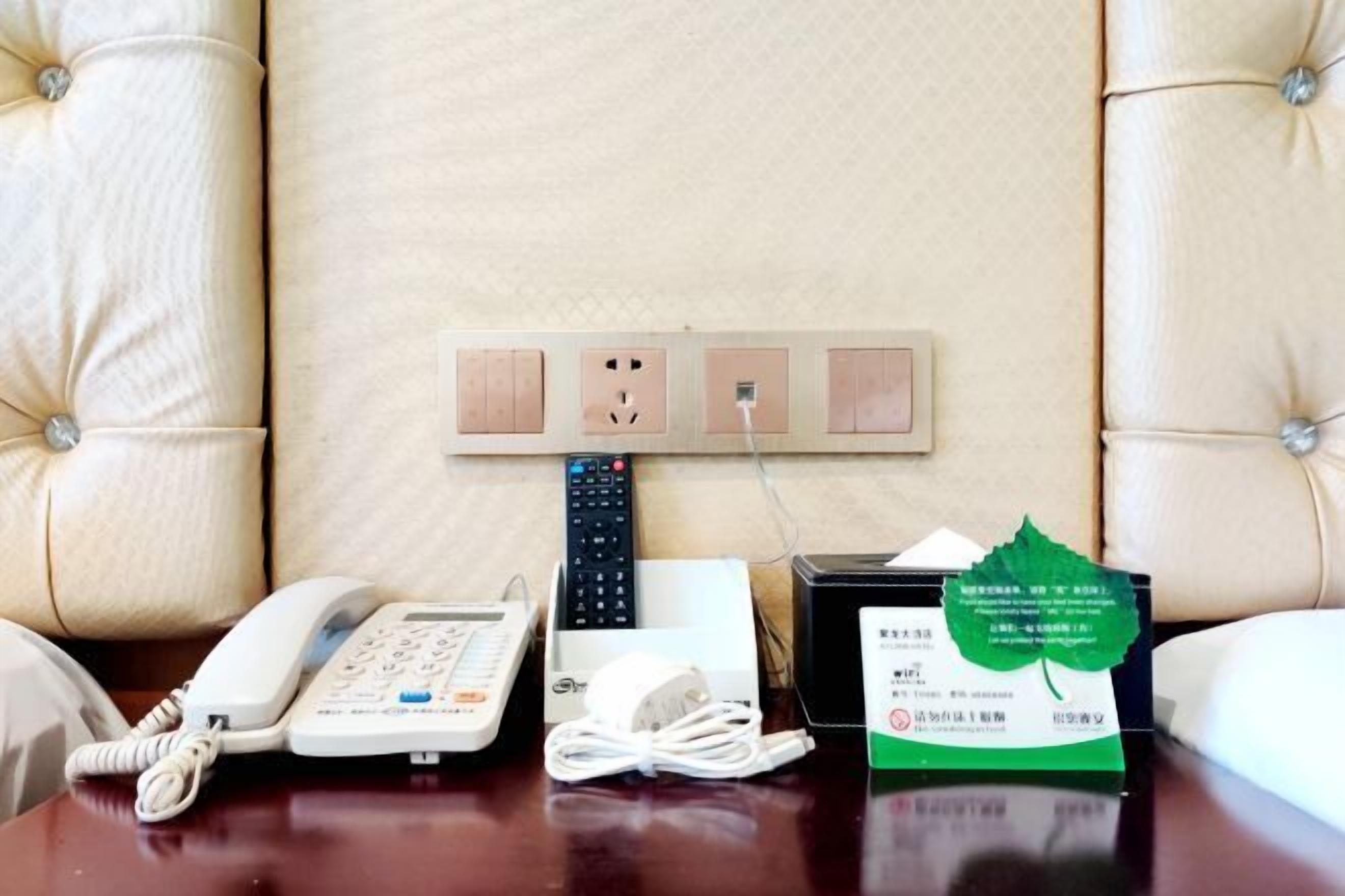 A hotel room with a tufted headboard, a telephone, a remote control, a power strip, and a green informational card on the table.