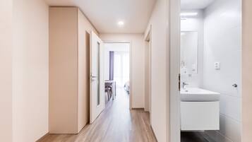 A hotel corridor with a wooden floor, white walls, and a bathroom with a sink and mirror.