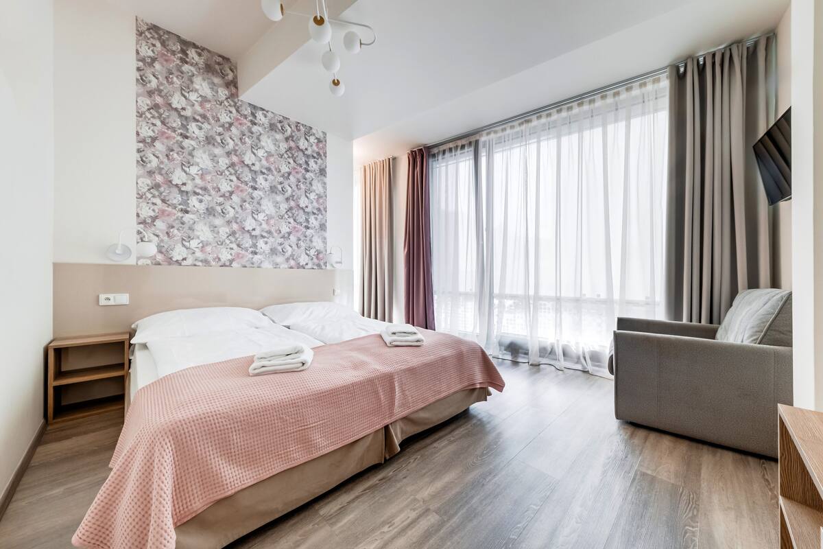Ein modernes Hotelzimmer mit Bett, einer Couch, einem großen Fenster mit Vorhängen und einem an der Wand befestigten Fernseher.