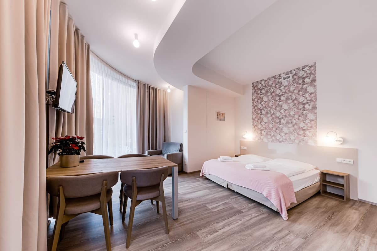 Ein modernes Hotelzimmer mit gewölbter Decke, einem Bett mit rosa Decke, einem Esstisch mit Stühlen und einem an der Wand befestigten Fernseher.