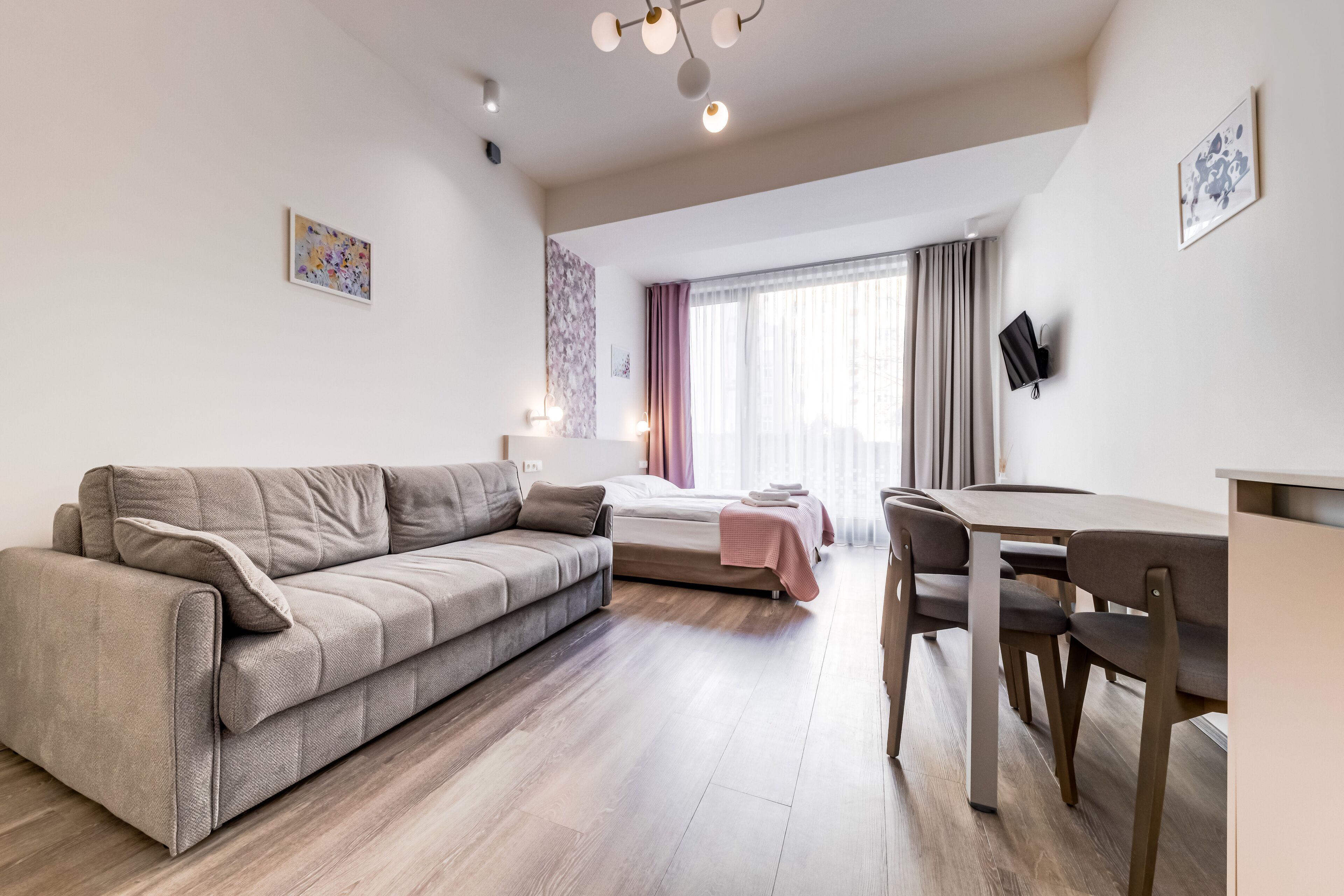 A modern living room with a sofa, a dining table with chairs, a bed with a pink blanket, and a TV mounted on the wall.