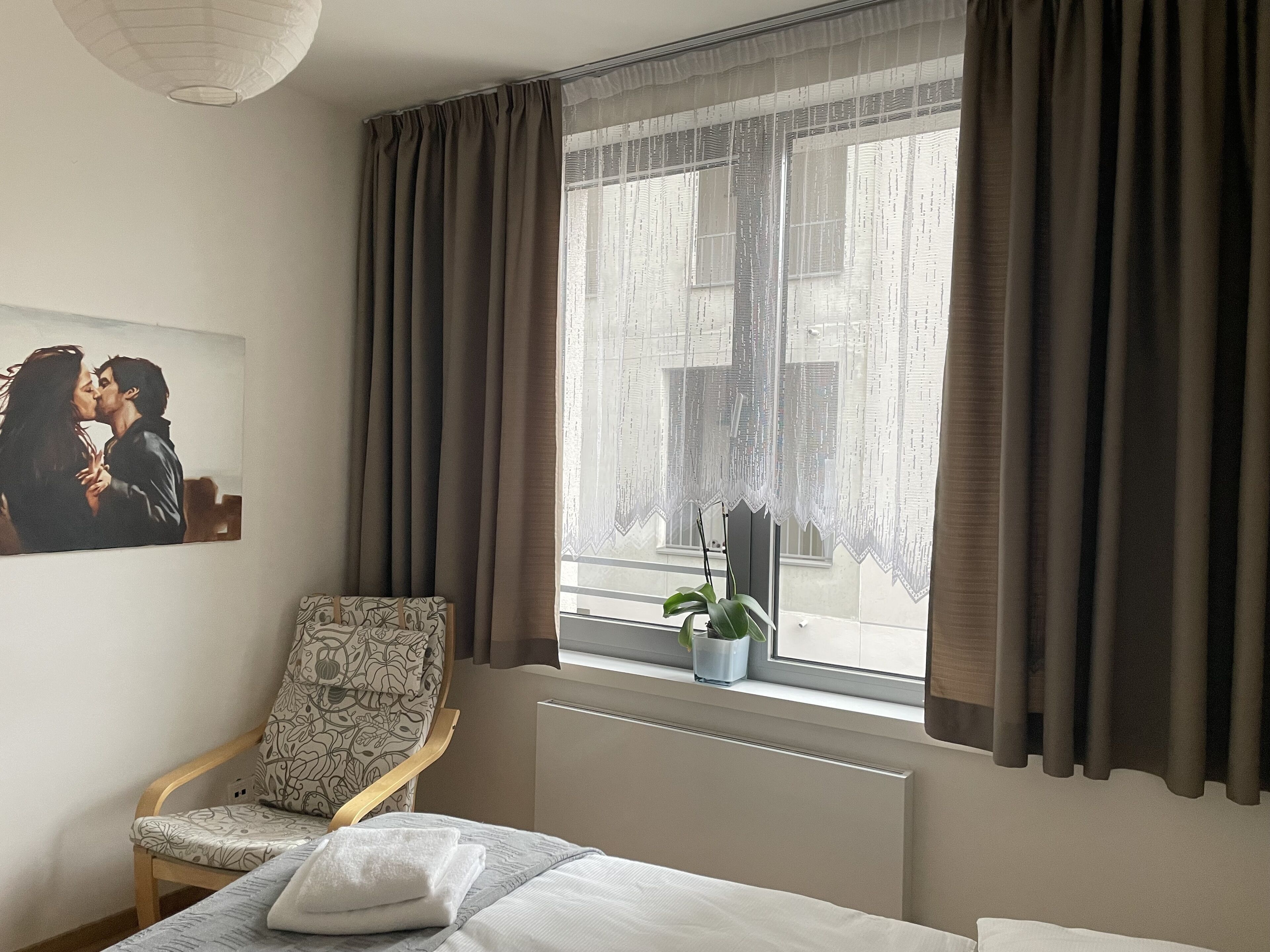 Habitación de hotel con cama, una silla, ventana con cortinas, radiador y una planta en maceta.