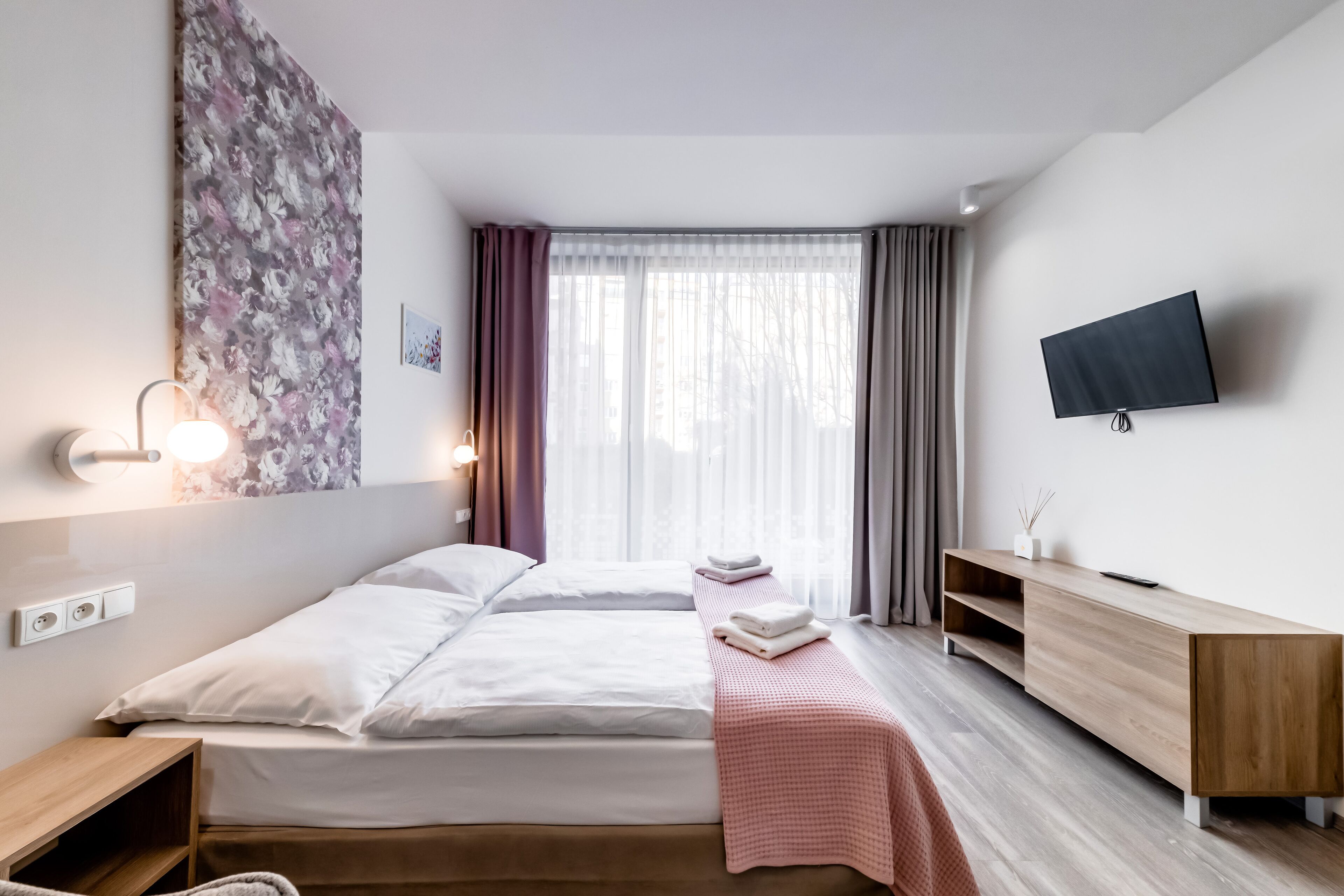 A modern hotel room with a bed, bedside table, TV, and a large window with curtains.