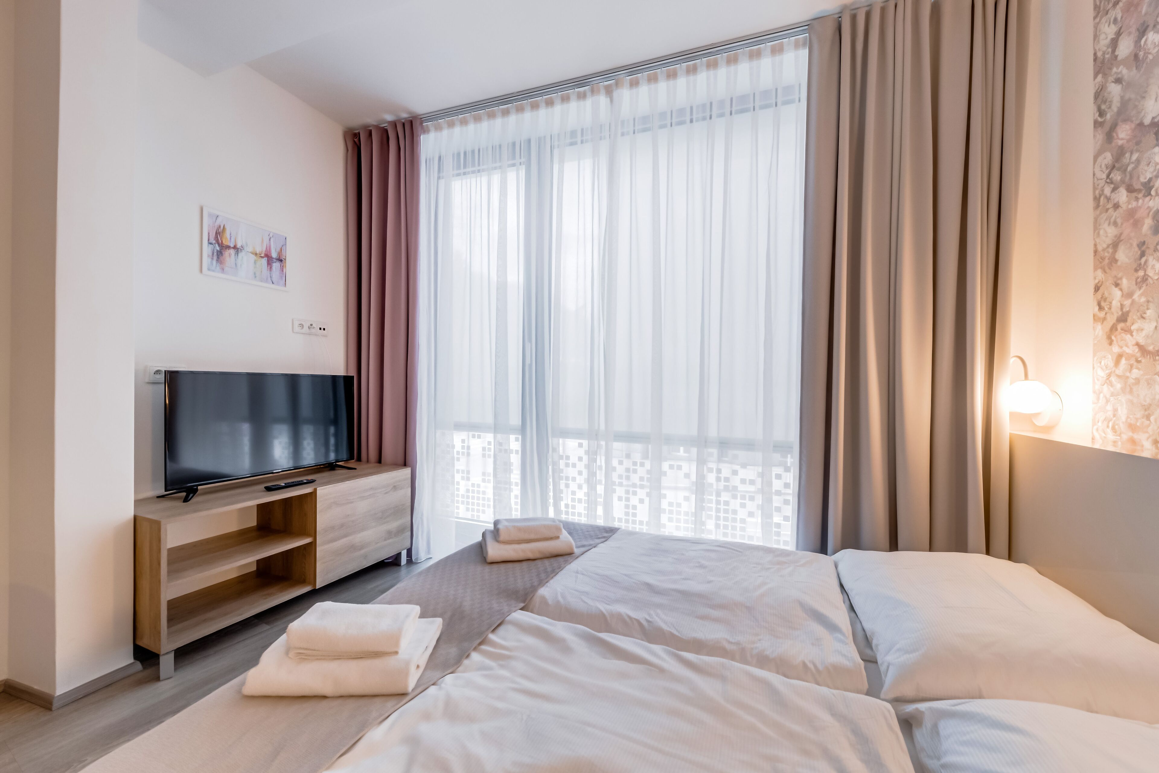 A hotel room with a TV on a wooden stand, a bed with white linens, a small table with towels, and a window with sheer curtains.