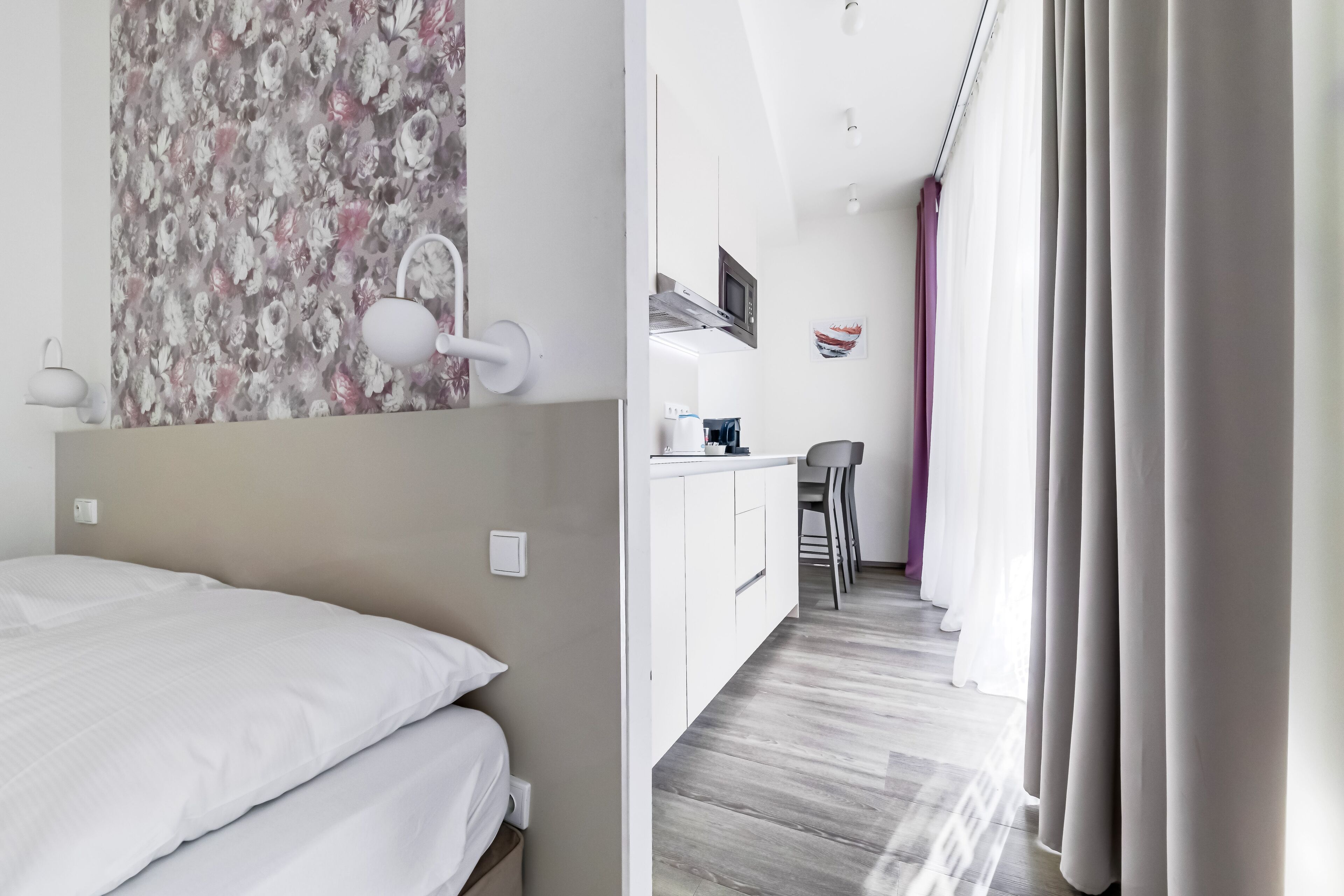 A hotel room with a bed, a small kitchenette, and a view of the hallway.