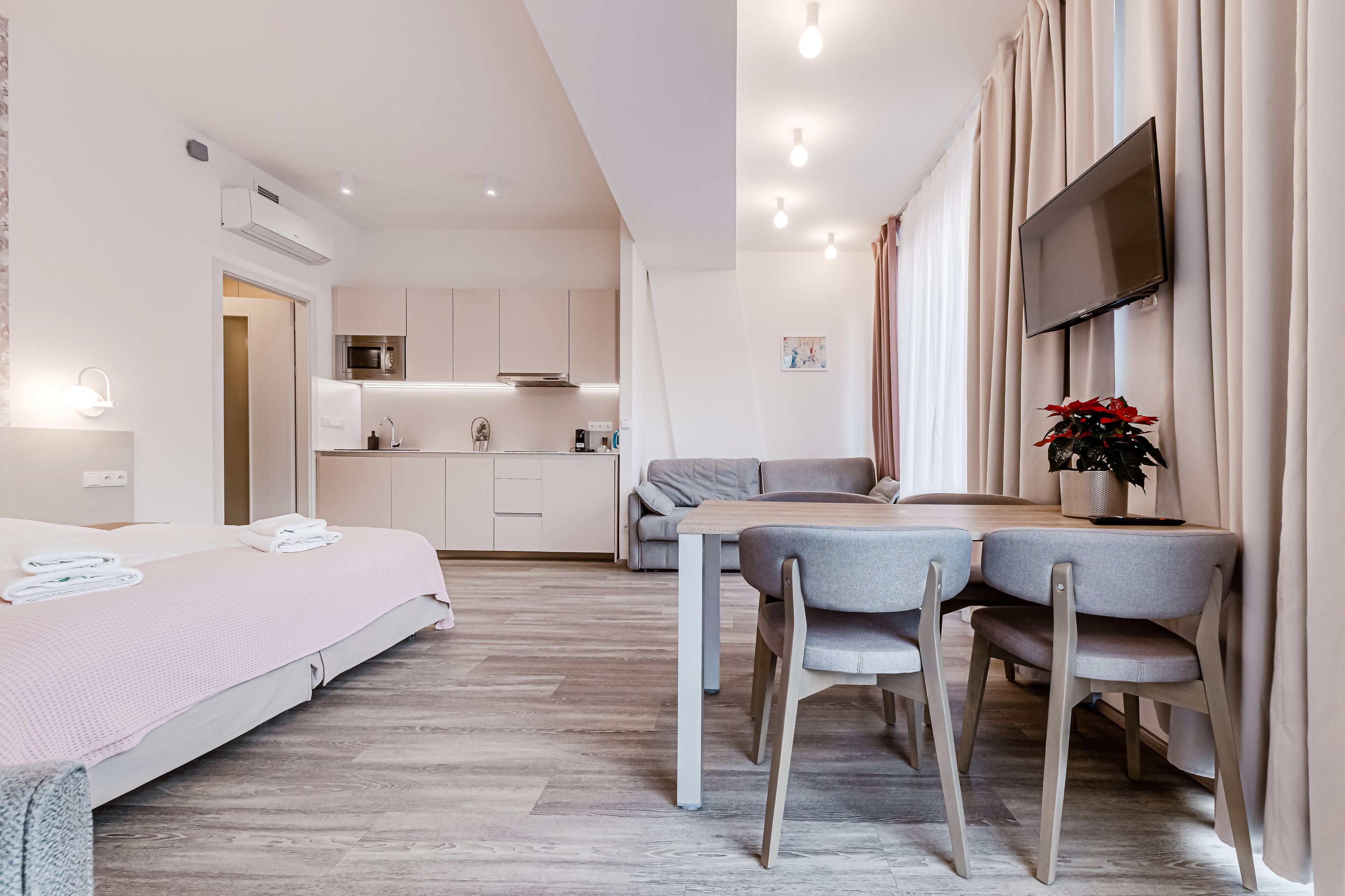 Et moderne hotelværelse med seng, spiseplads med stole, et fjernsyn ophængt på væggen og et køkkenområde i baggrunden.
