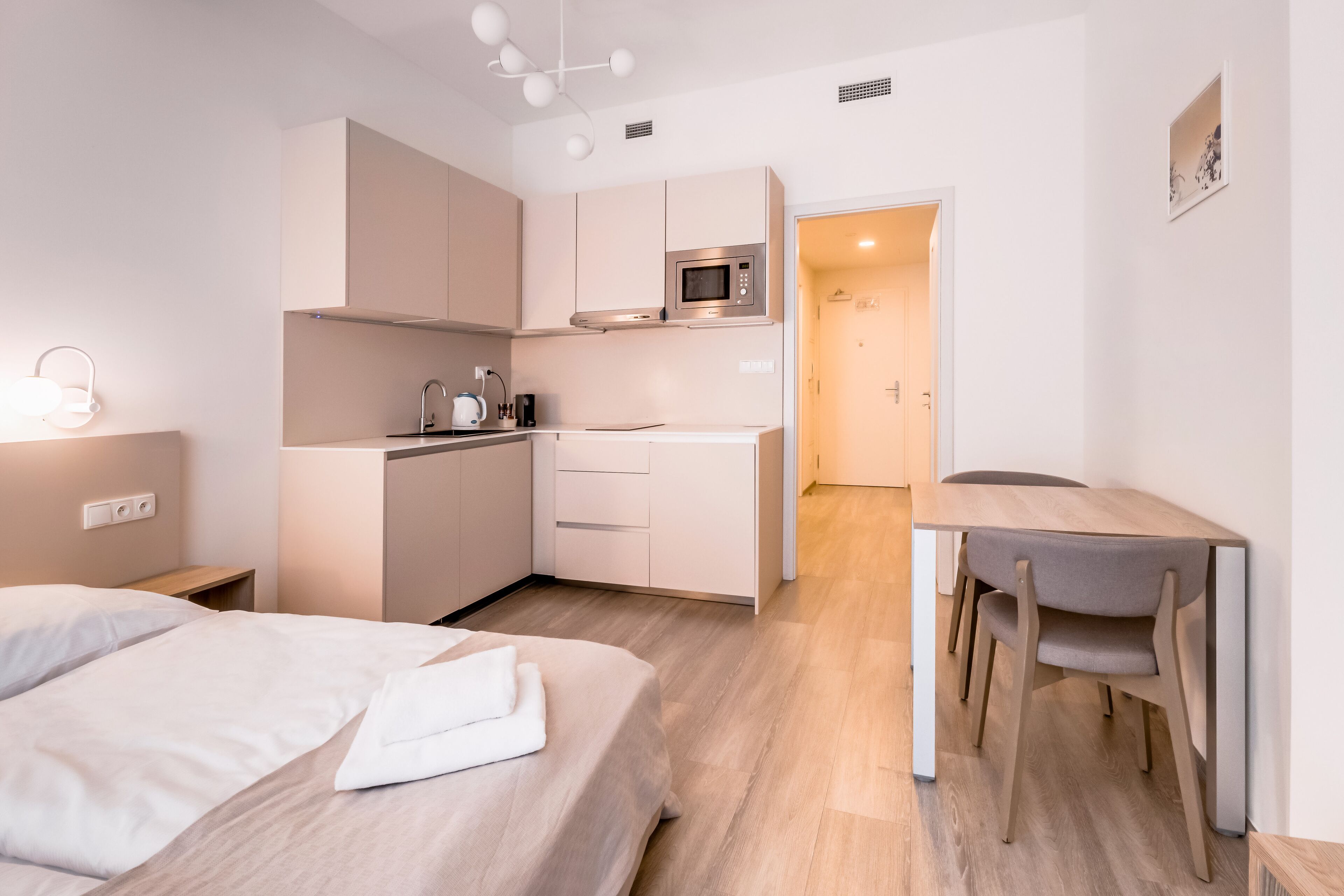 Quarto de hotel moderno com cama, pequena cozinha, mesa de jantar com cadeiras e banheiro visível por uma porta aberta.