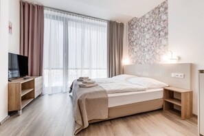 A modern hotel room with a bed, a TV, a wooden nightstand, and a large window with curtains. - KORA APARTMENTS (Prague)
