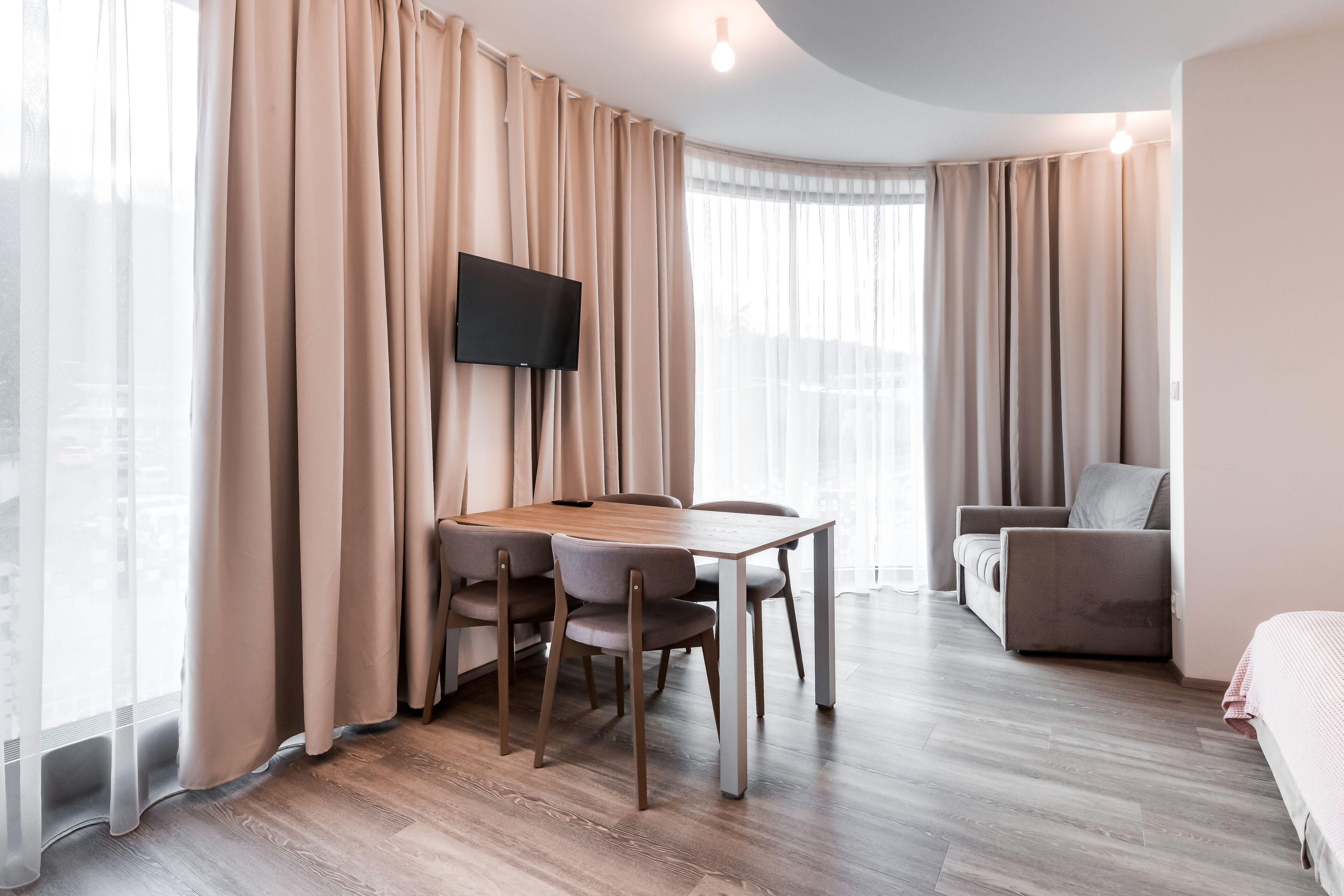 A hotel room with a wooden dining table, four chairs, a flat-screen TV mounted on the wall, a sofa, and a bed.