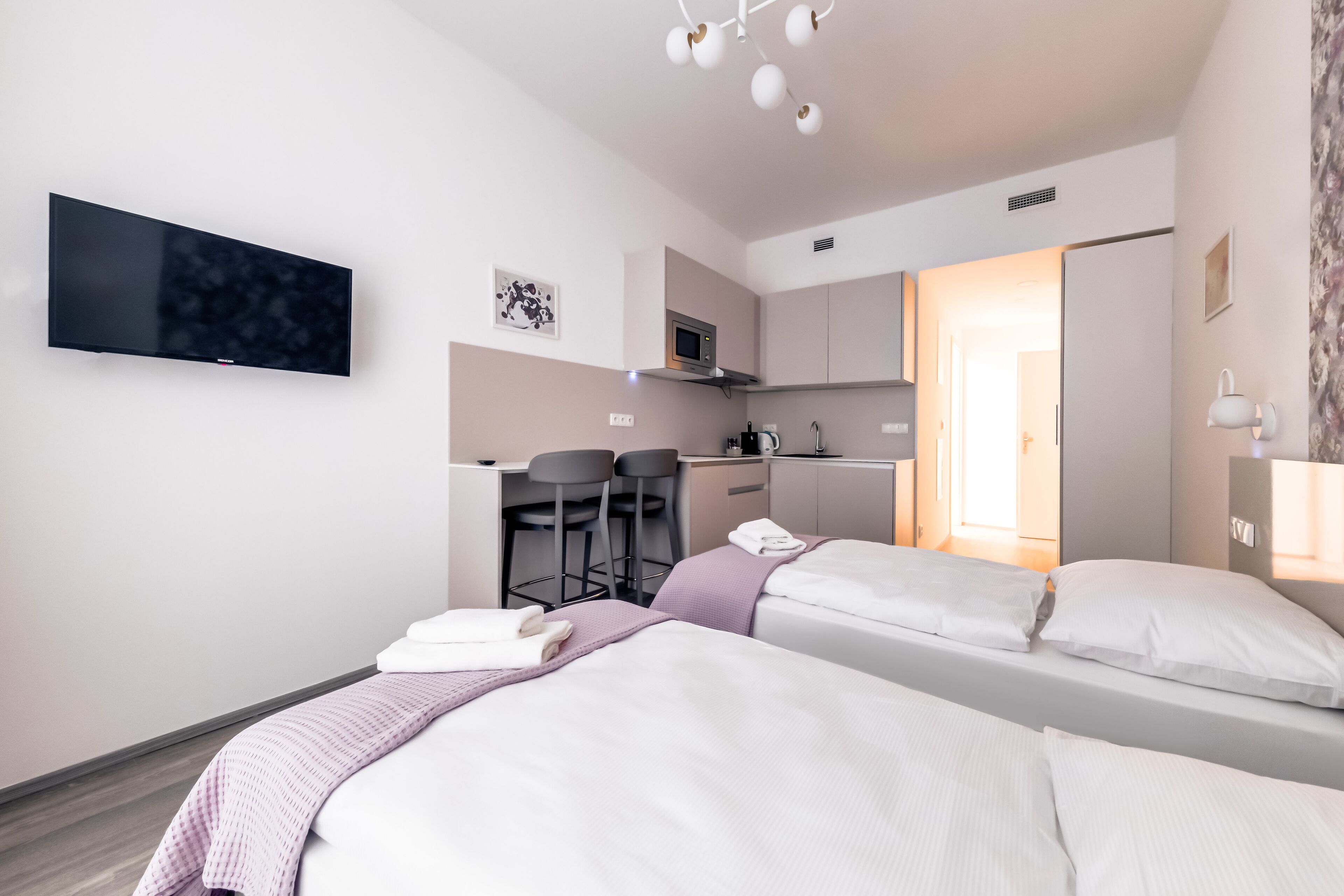 A modern hotel room with two beds, a flat-screen TV, a kitchenette, and a bathroom visible through an open door.