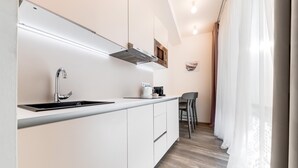 A modern kitchen with white cabinetry, a stainless steel sink, and a built-in range hood. There is a dining area with a table and chairs, and a window with sheer curtains. - KORA APARTMENTS (Prague)