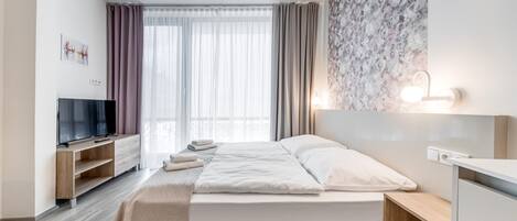 Una habitación de hotel moderna con cama, televisor, escritorio y un amplio ventanal con cortinas.