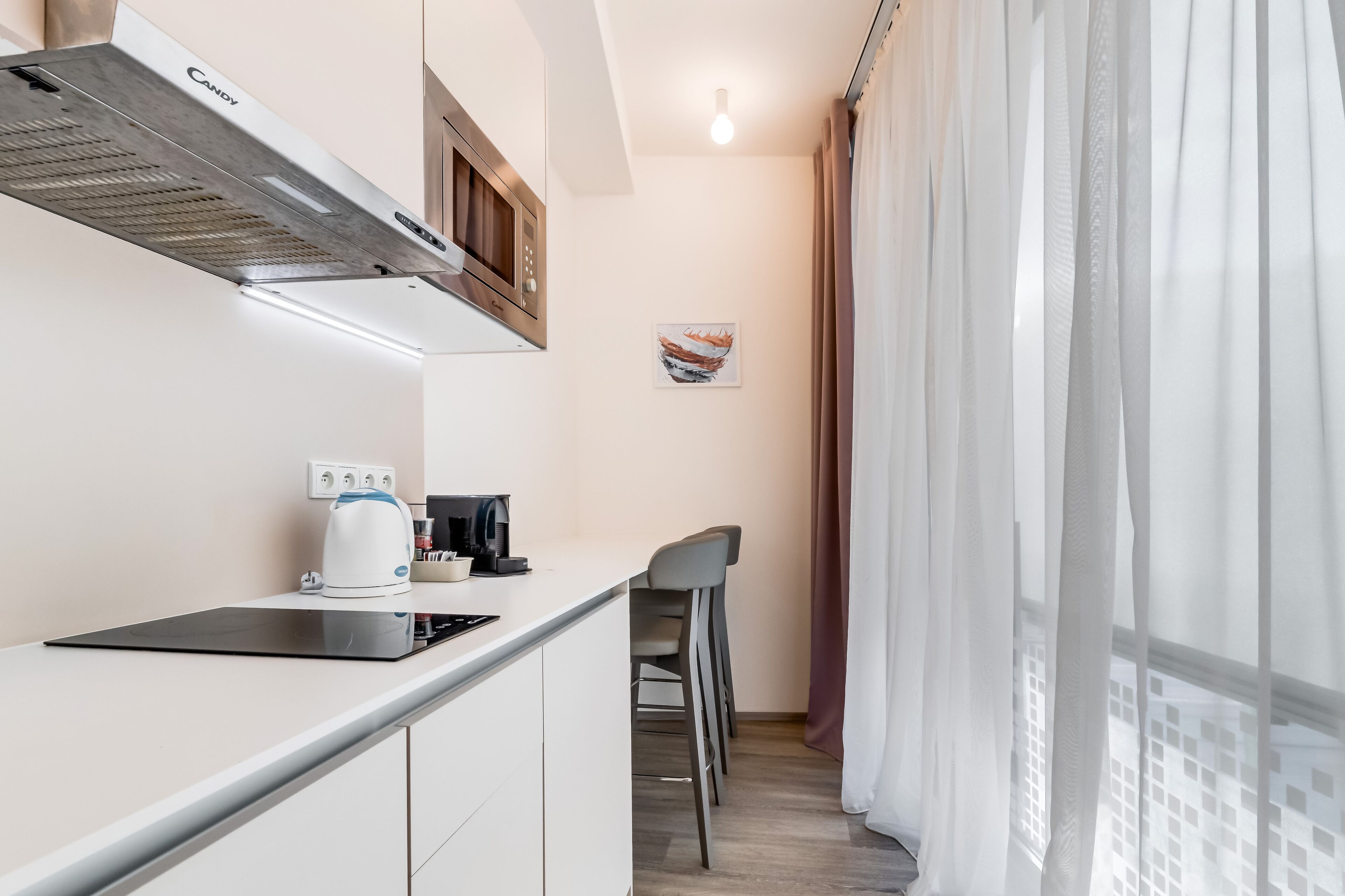 A modern kitchen with a built-in stove, a coffee maker, and a microwave. There are two bar stools and a window with sheer curtains.
