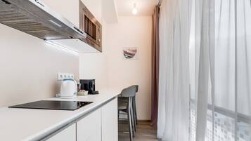 A modern kitchen with a built-in stove, a coffee maker, and a microwave. There are two bar stools and a window with sheer curtains.