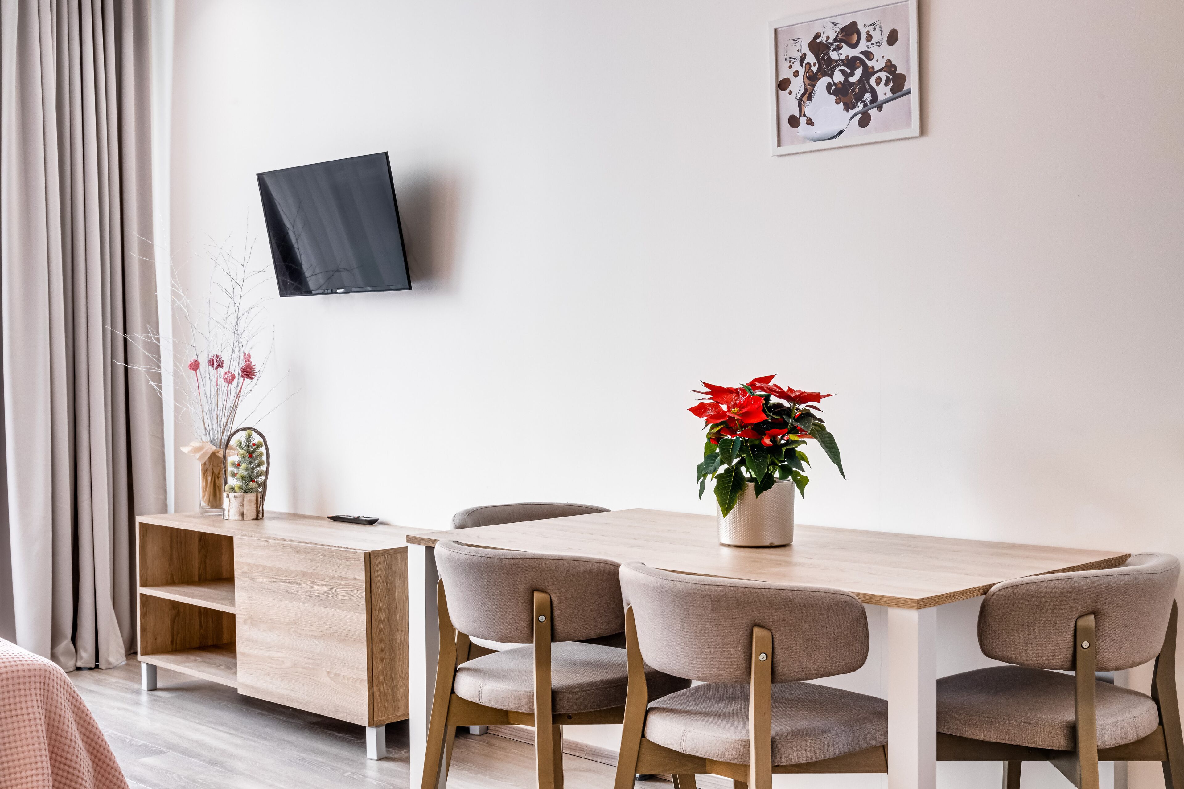 A modern dining area with a wooden table and four chairs, a TV mounted on the wall, and a potted plant with red flowers.