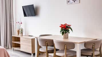 A modern dining area with a wooden table and four chairs, a TV mounted on the wall, and a potted plant with red flowers.