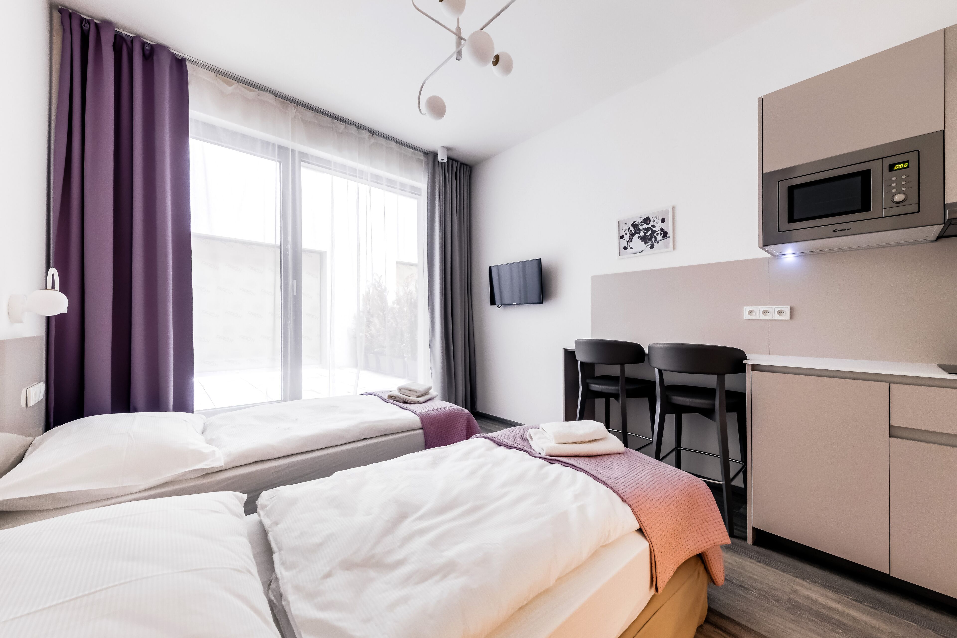 A hotel room with two beds, a kitchenette, a TV, and a window with curtains.