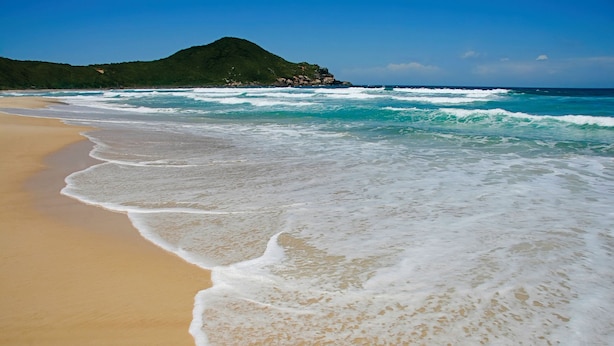 A beach with waves gently lapping onto the shore, a clear sky, and a green hill in the background. - Pousada Vigia das Marés - Praia do Rosa - SC (Imbituba)