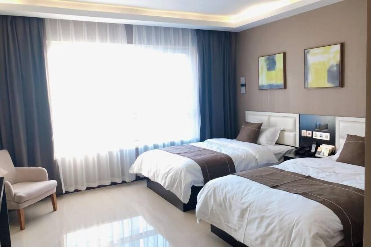 A hotel room with two beds, a chair, a large window with curtains, and artwork on the wall.