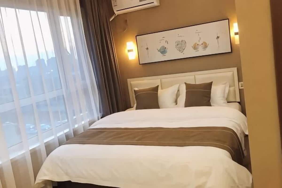 A hotel room with a large bed, a headboard with multiple pillows, a window with curtains, and a wall-mounted artwork.