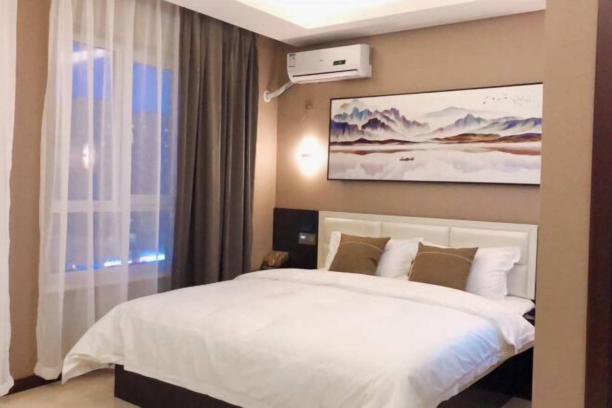 A hotel room with a bed, an air conditioning unit, a painting, and a window with curtains.