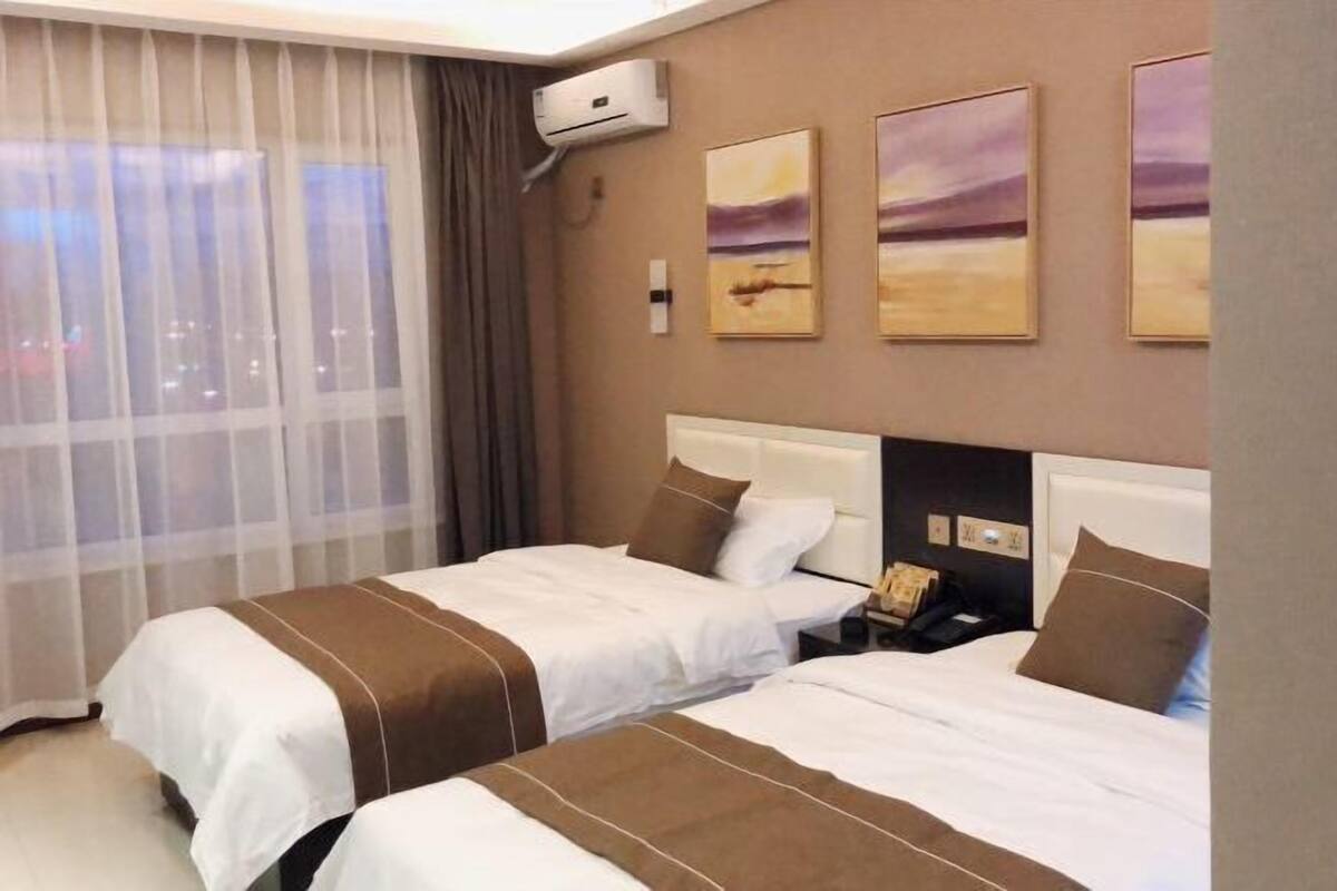 A hotel room with two beds, a window with sheer curtains, an air conditioning unit, and three landscape paintings on the wall.