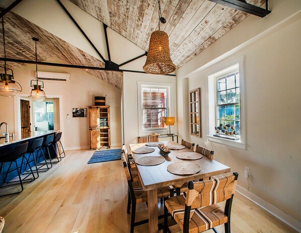 A spacious dining area with a wooden table and chairs, a hanging woven light fixture, and a view of the outdoors through a window.