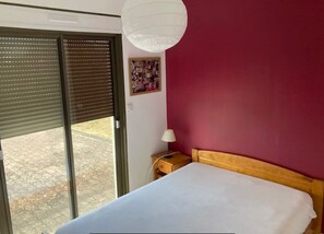 A bedroom with a bed, a nightstand, a lamp, a window with blinds, and a wall-mounted light fixture. - House Frehel-Plage (Fréhel)