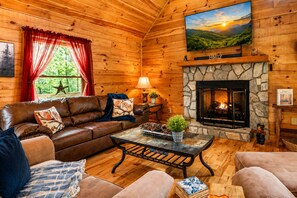 A cozy log cabin interior with a stone fireplace, leather sofa, and a television mounted on the wall.