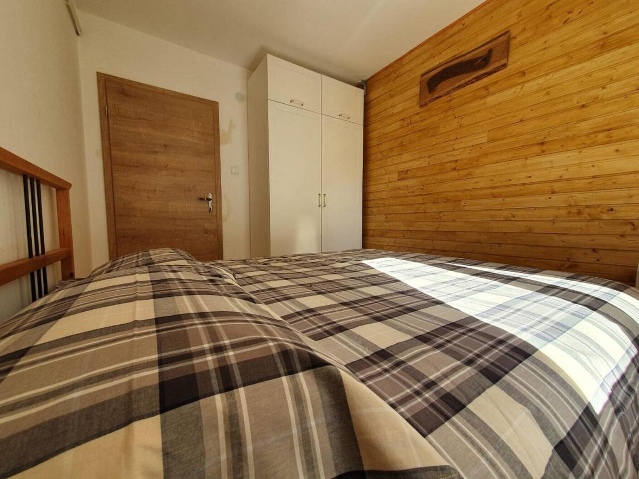 A bedroom with a wooden headboard, a plaid bedspread, a wardrobe, and a wooden wall panel.