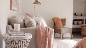 Ein gemütliches Wohnzimmer mit einer Couch, einem runden Beistelltisch mit Buch und Kerze, einem Sessel mit Decke und einem Bücherregal im Hintergrund.