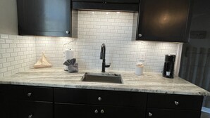 Uma cozinha com armários escuros, painel de azulejos branco, pia e máquina de café.
