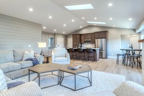 A spacious living area with a sofa, coffee table, and a dining table with chairs. A kitchen with modern appliances and bar stools is visible in the background.