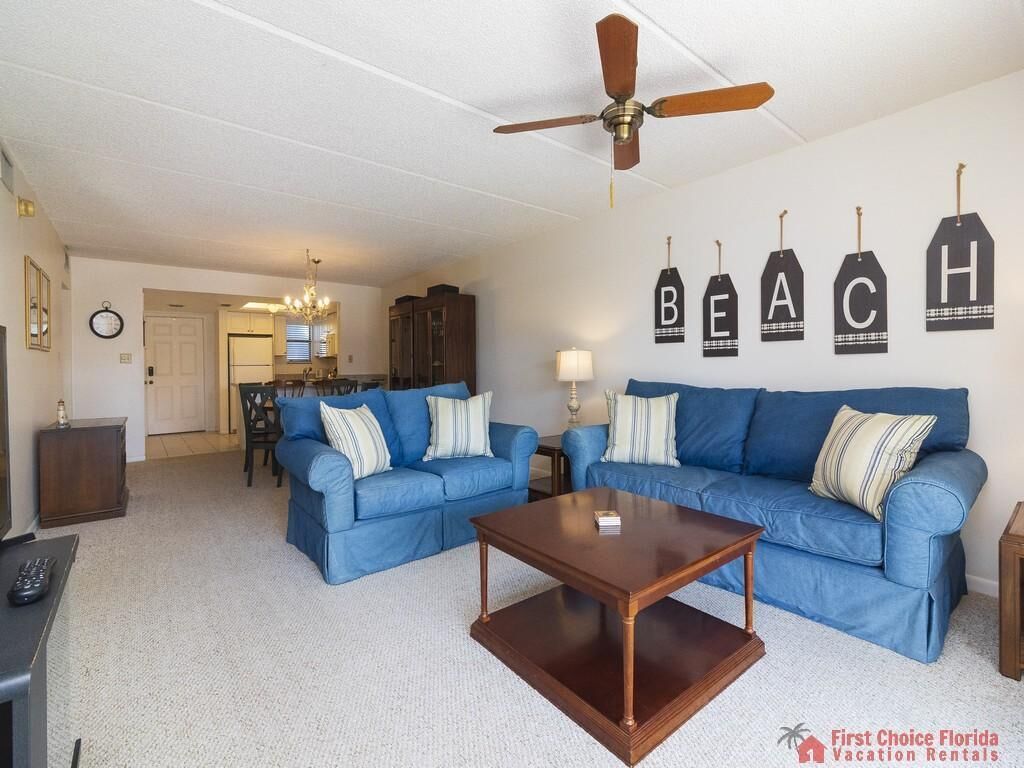 Ein Wohnzimmer mit zwei blauen Sofas, einem Holztisch, einem Deckenventilator und Wanddekorationen mit der Aufschrift "BEACH".