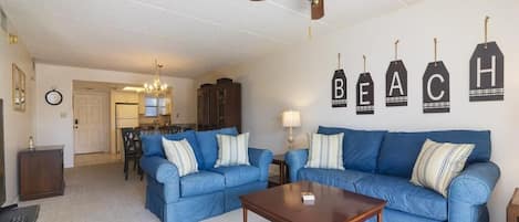 A living room with two blue sofas, a wooden coffee table, a ceiling fan, and wall decorations spelling out "BEACH".