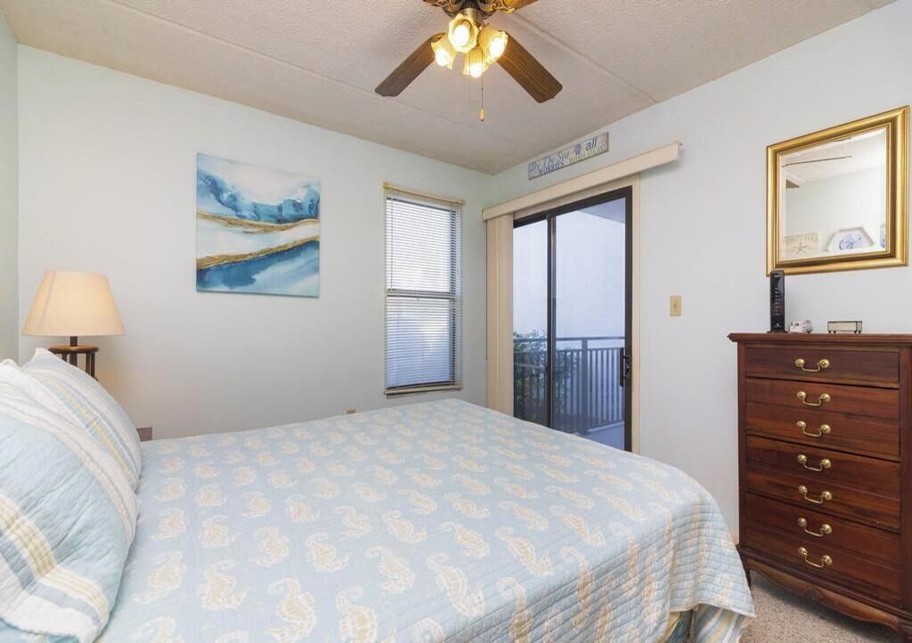 A bedroom with a bed, a dresser, a ceiling fan, a window with blinds, a painting, and a balcony door.