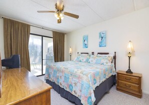 A hotel room with a bed, a desk, a chair, a ceiling fan, two wall-mounted lamps, and a window with curtains. - Excellent Beach Location ~ Pool ~ Great Price! (Saint Augustine)