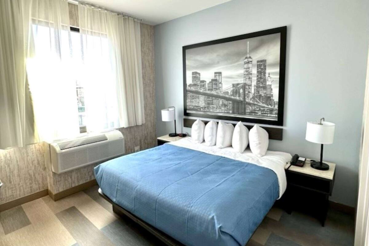 A hotel room with a bed, a cityscape artwork on the wall, a window with curtains, and a bedside table with a lamp.