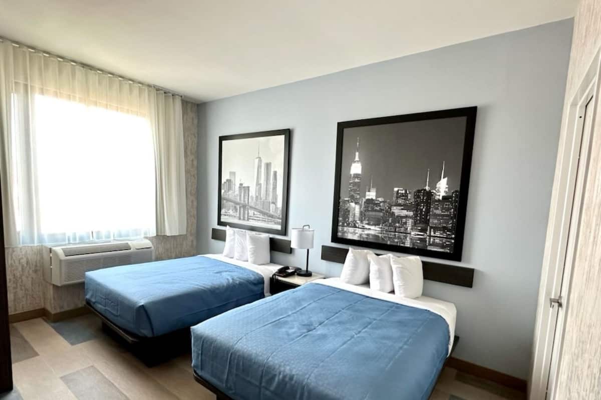 A hotel room with two beds, a cityscape artwork on the wall, a window with curtains, and a bedside table with a lamp.
