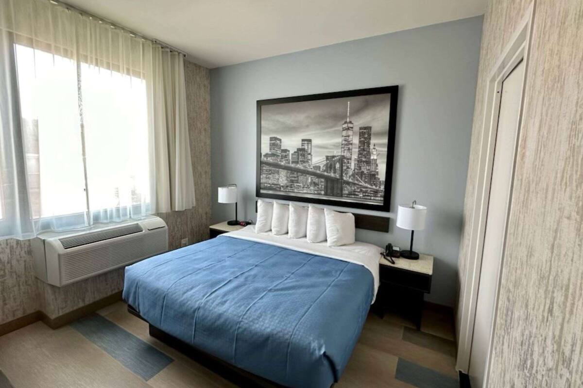 A hotel room with a large bed, a cityscape artwork on the wall, a window with curtains, and a bedside table with a lamp.