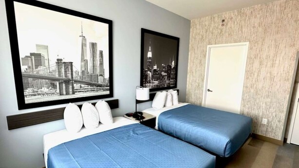 Ein Hotelzimmer mit zwei Betten, Blick auf die Stadt, einer Tür und einem Nachttisch mit einer Lampe.