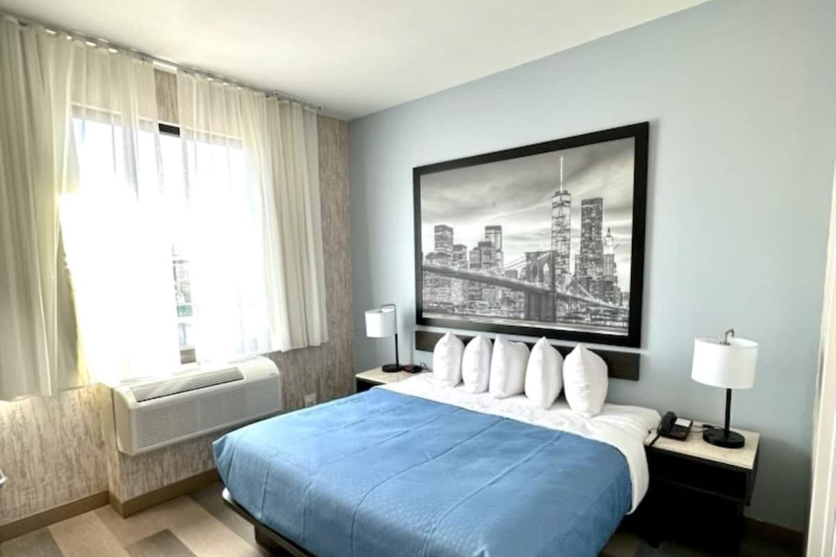 A hotel room with a bed, a window with curtains, a wall-mounted picture, bedside lamps, and a nightstand.