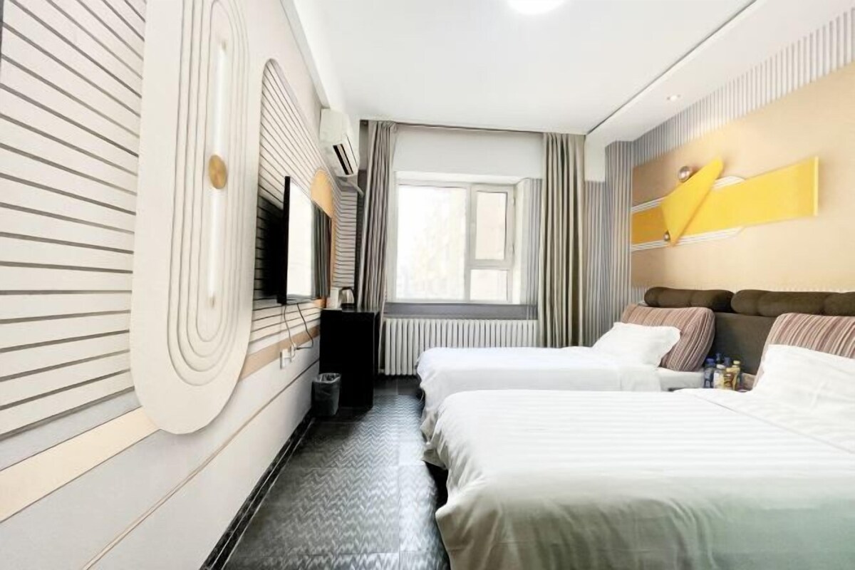 A hotel room with two beds, a large window, a radiator, and a wall-mounted TV.